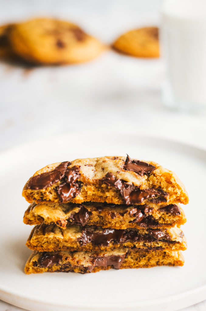 Stack of halved chocolate chip cookies with milk and whole cookies in the rear