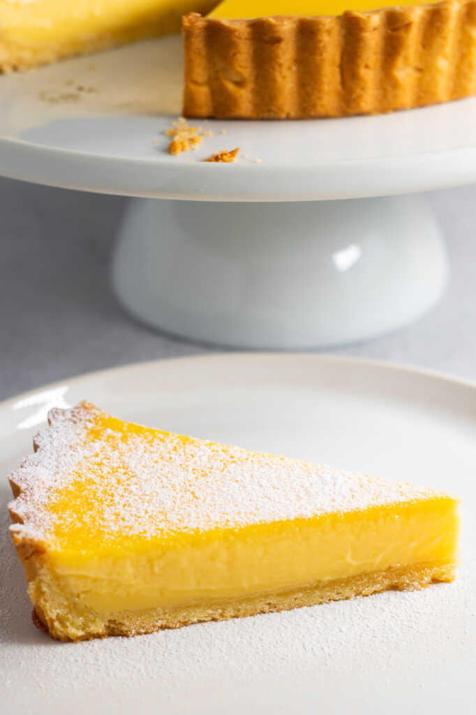 Slice of no-bake lemon curd tart dusted with icing sugar on a white plate, with the whole tart in the background.