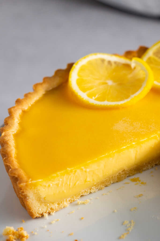 Close-up of a creamy no-bake lemon curd tart slice with buttery pâte sablée crust and lemon slices on top.