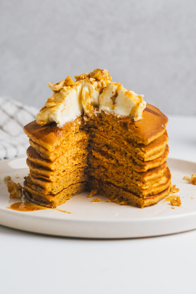 Stack of carrot cake pancakes topped with cream cheese frosting and walnuts cut open to reveal centre