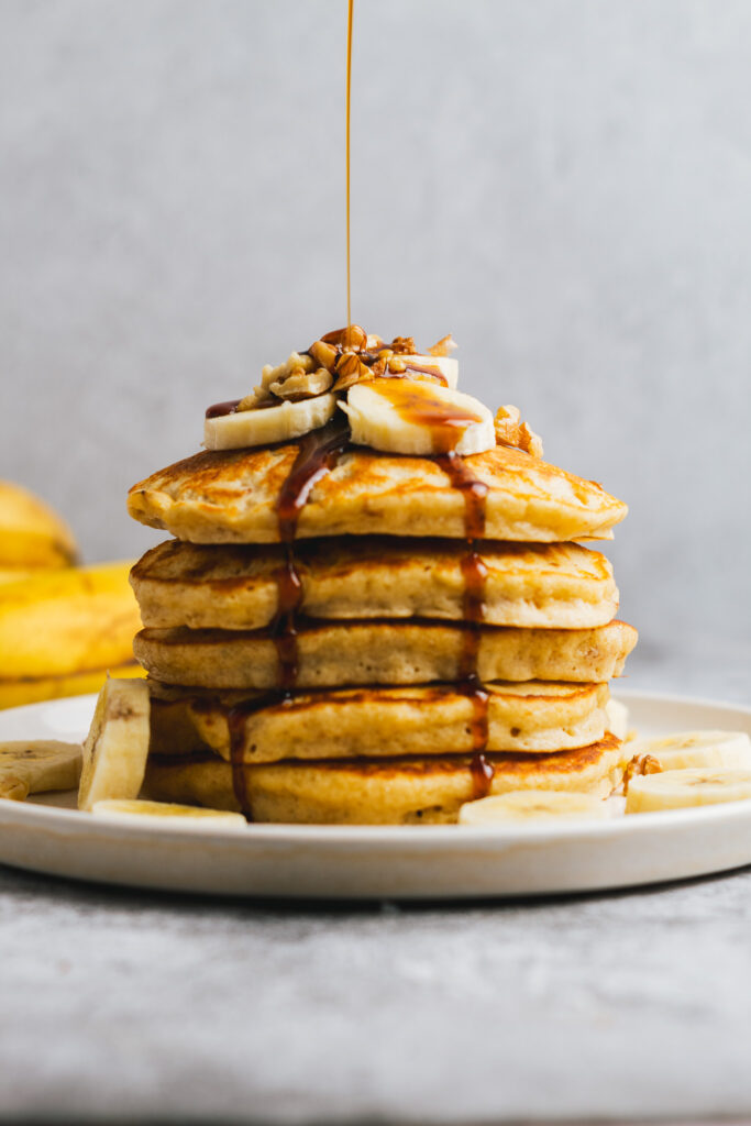 Stack of Banana Walnut pancakes topped with bananas and chopped walnuts, drizzled with maple syrup