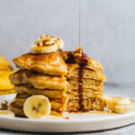 Stack of banana walnut pancakes with section cut out, drizled with maple syrup
