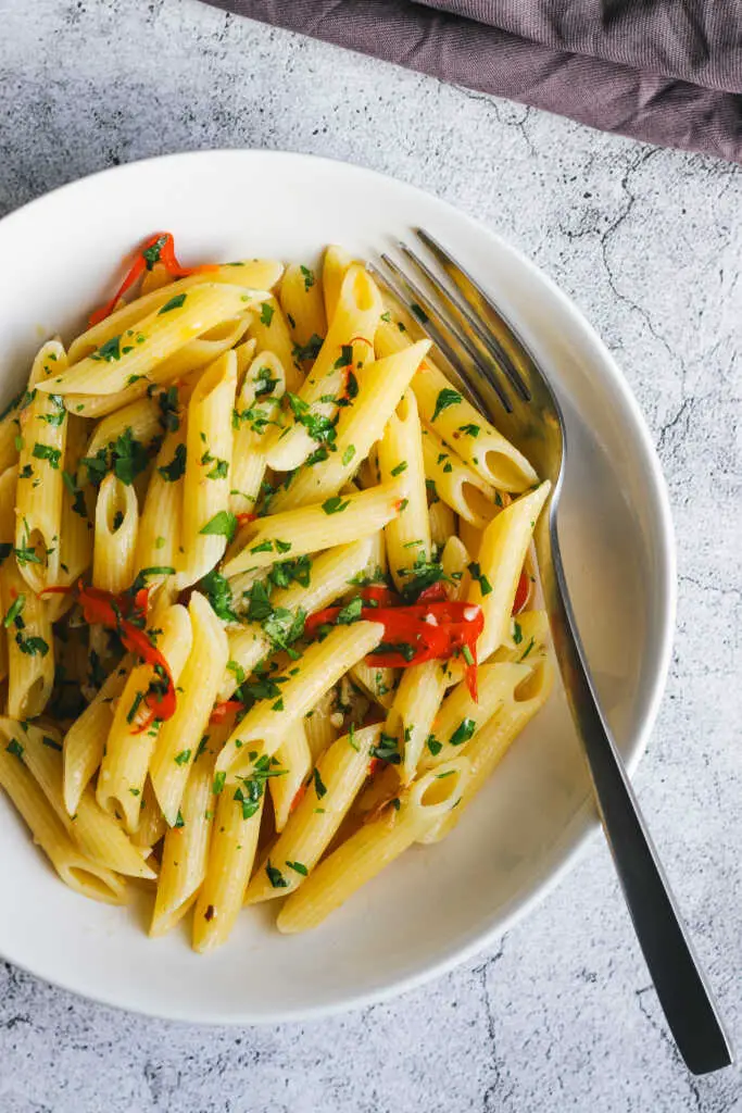 Overhead bowl of Penne Aglio e Olio with garlic, chilli and parsley on a grey background