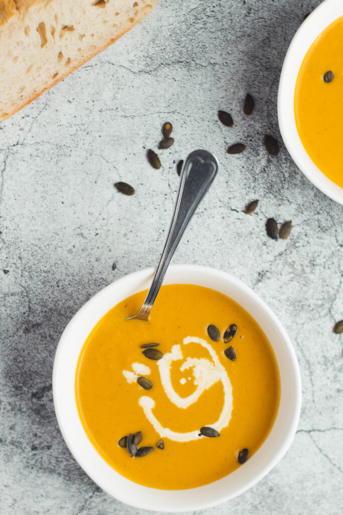 Two bowls of carrot ginger pumpkin soup with some bread and scattered pumpkin seeds