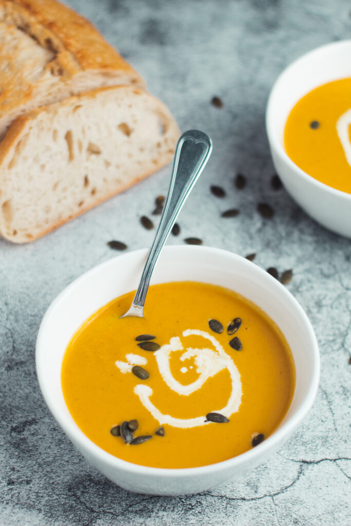 Two bowls of carrot ginger pumpkin soup with some bread and scattered pumpkin seeds