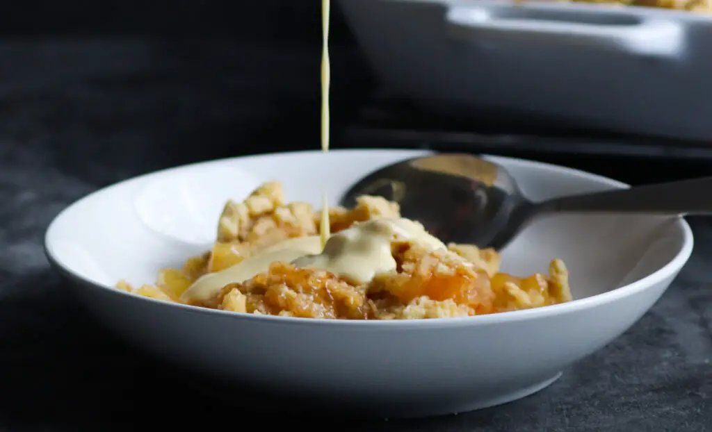 bowl of apple crumble with custard being poured into bowl