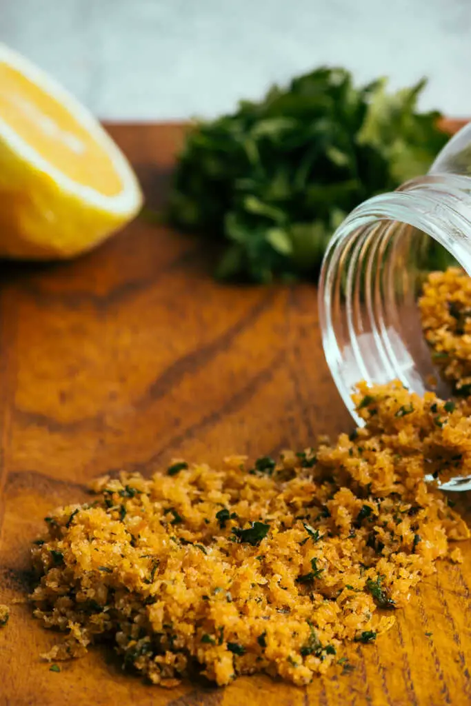 A jar of pangrattato breadcrumbs on a chopping board with lemon and parsley