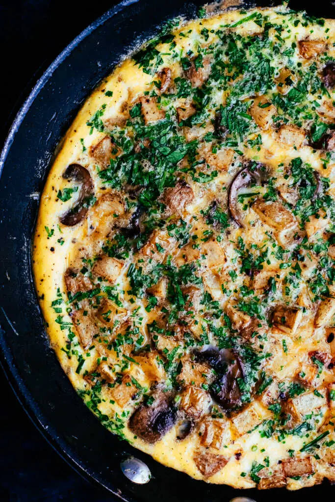Close-up of a rustic country omelette with crispy potatoes, mushrooms, bacon, and fresh herbs in a frying pan