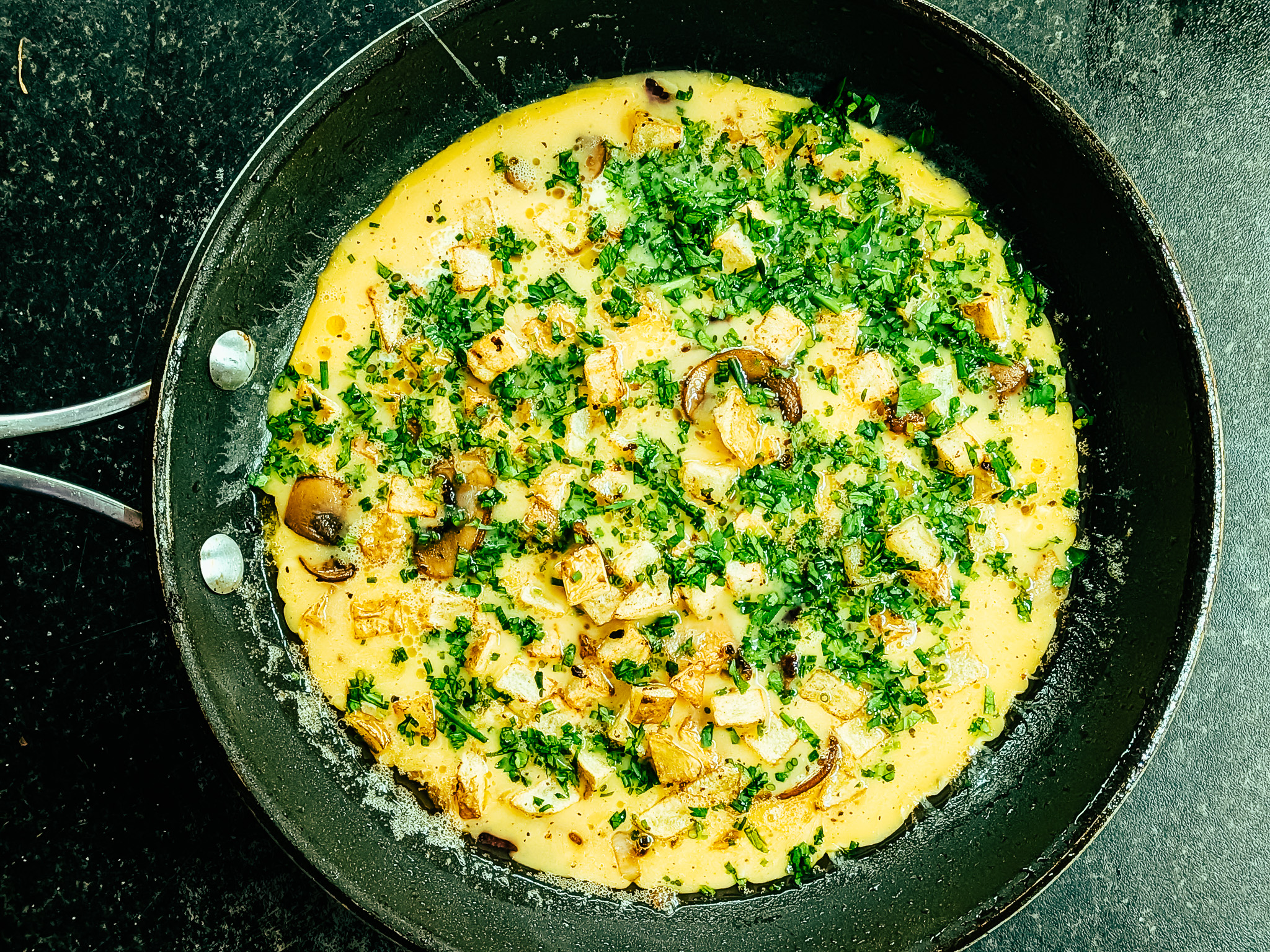 Country omelette cooking in a skillet with diced potatoes, mushrooms, and fresh herbs