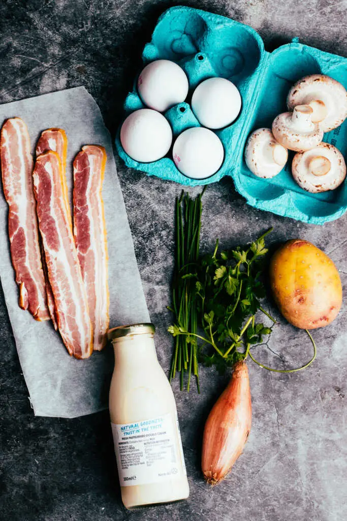 Ingredients for a rustic country omelette with bacon, eggs, mushrooms, potato, herbs, and cream on a dark background