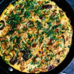 Country omelette with crispy potatoes, bacon, and mushrooms cooked in a skillet and topped with fresh herbs