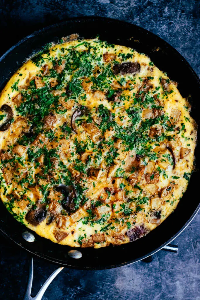 Country omelette with crispy potatoes, bacon, and mushrooms cooked in a skillet and topped with fresh herbs