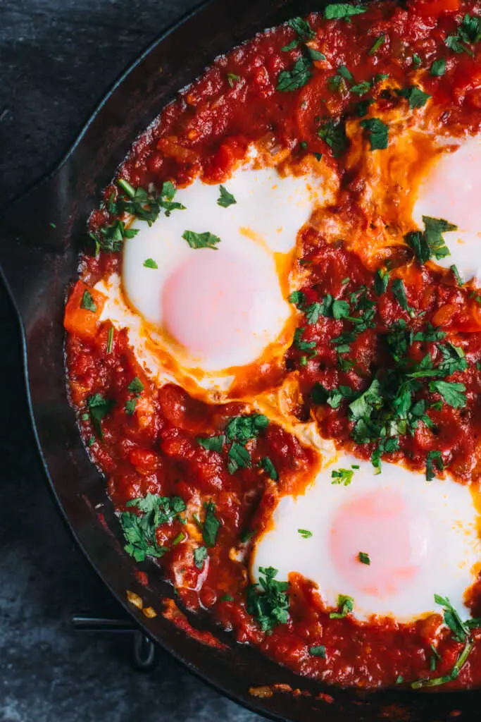 Close-up of Moroccan Shakshuka with soft-poached eggs in a rich rose-harissa tomato sauce topped with fresh herbs.